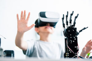 Waist up of a young smart boy wearing VR glasses and controlling his robotic hand